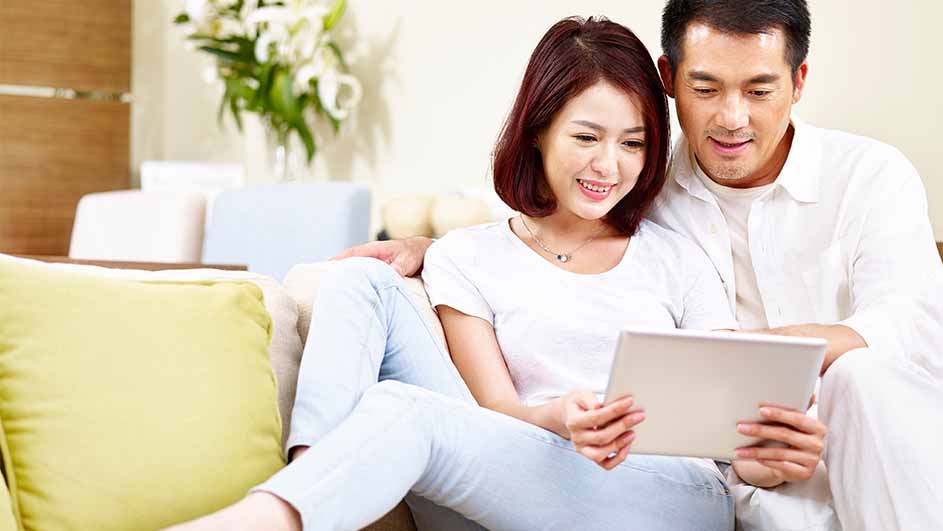 Een vrouw en een man zitten op een bank en kijken naar een tablet. Er staat een vaas met bloemen op de achtergrond.