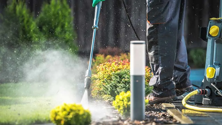 Persoon die een tuinpad schoonmaakt met een hogedrukreiniger naast planten en een lantaarnpaal.