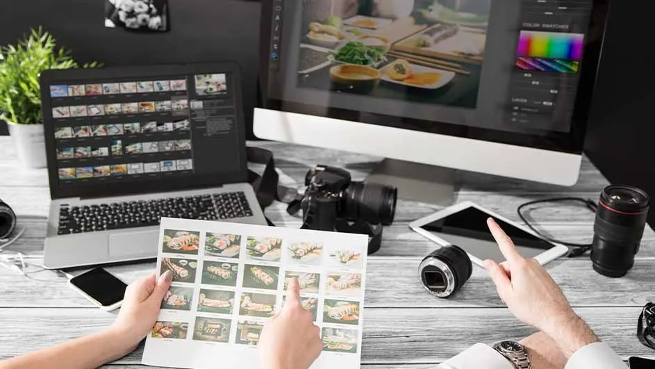 Desk setup with laptop, monitor, camera, tablet, and lenses. Hands point to images of food on printed paper and tablet screen.