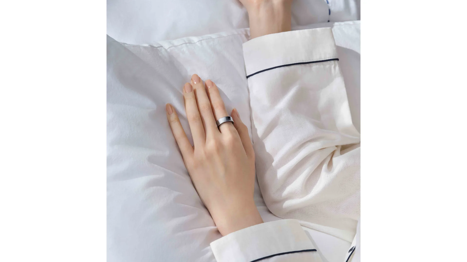 Hand met een zilveren ring ligt op een wit kussen, gekleed in een witte pyjama met donkere rand.