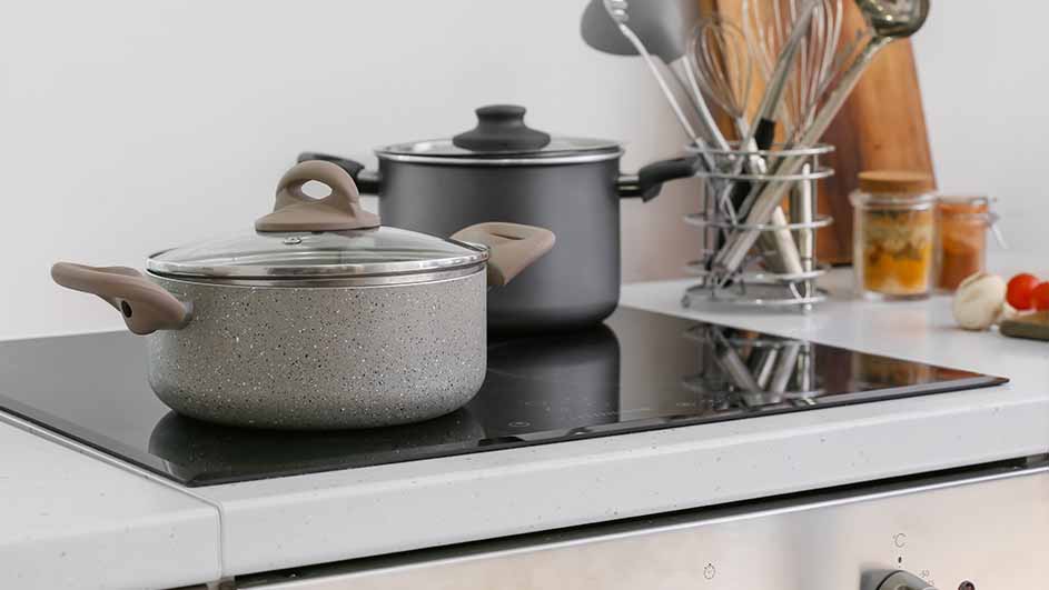 Twee pannen staan op een zwarte kookplaat. Een rek met keukengerei, kruidenpotjes en groenten staan op het aanrecht.