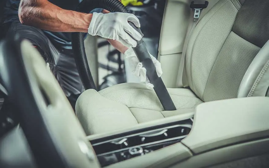 Een persoon met witte handschoenen stofzuigt een lichtgekleurde autostoel grondig voor een perfect gereinigd auto-interieur.