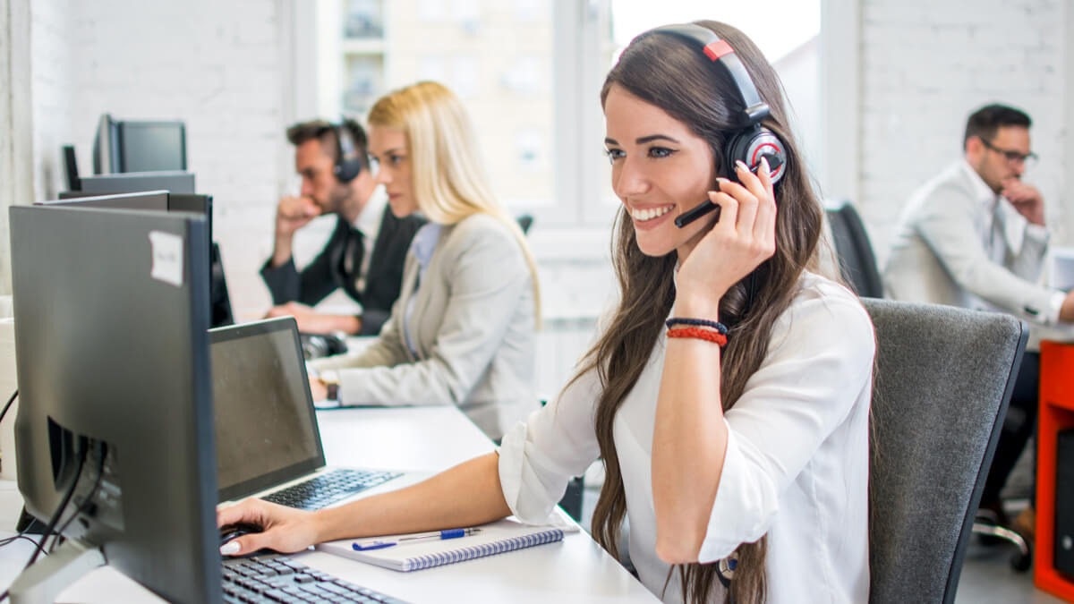 Office scene with people working at computers, wearing headsets. A woman smiles, using a computer and speaking into the headset.