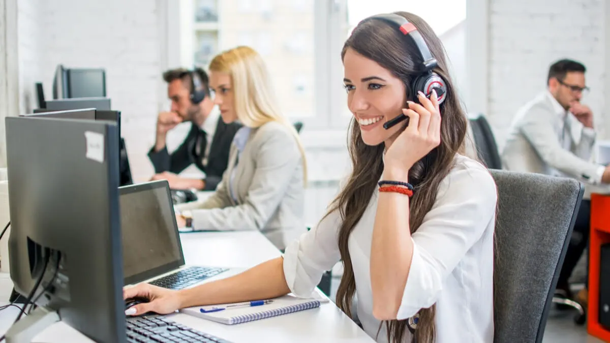 Een kantoor met medewerkers achter computers, headsets dragend. Een vrouw lacht en gebruikt een headset. Monitoren en bureaus zijn zichtbaar.
