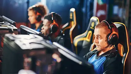 Drie mensen met headsets zitten aan computers in zwarte en gele gamingstoelen voor blauwe gordijnen.