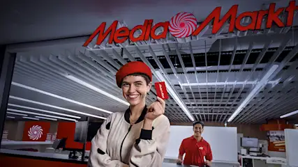 Een vrouw met een rode baret houdt een rode kaart vast onder een rood Media Markt logo in een winkel.