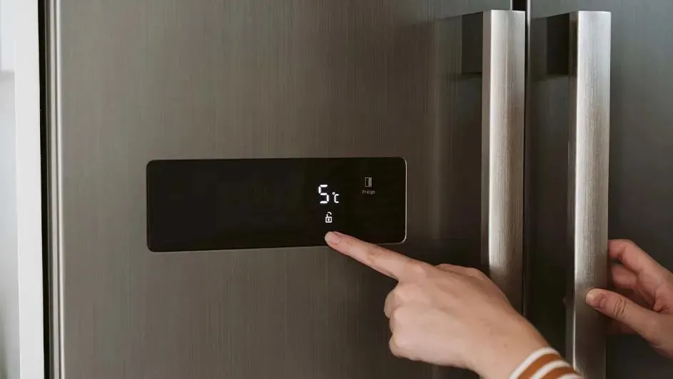 Hand uses the black control on a stainless fridge door to set 5°C, fingertip gently tapping the illuminated touch button.