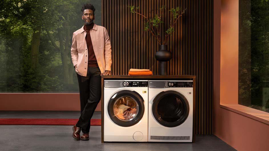 Een man leunt tegen een wasmachine en droger in een kamer met houten lambrisering en een raam met uitzicht op bomen.