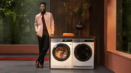 Een man leunt tegen een wasmachine en droger in een kamer met houten lambrisering en een raam met uitzicht op bomen.