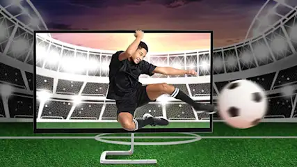 Een voetballer in zwart trapt krachtig op het veld; een grote bal vliegt uit het scherm in een fel verlicht stadion.
