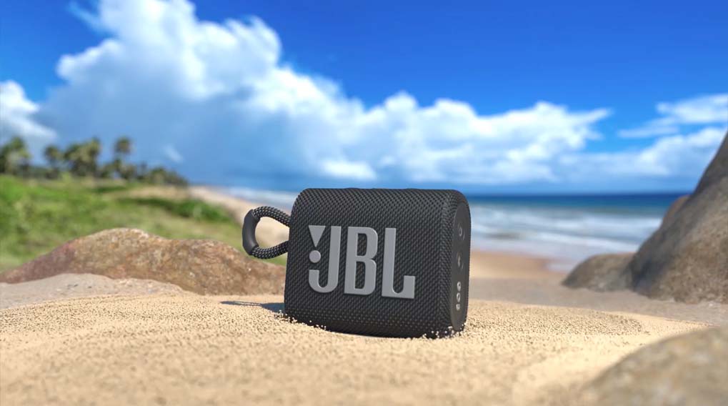 Een grijze JBL-speaker staat op een zandstrand met een blauwe lucht en zee op de achtergrond.