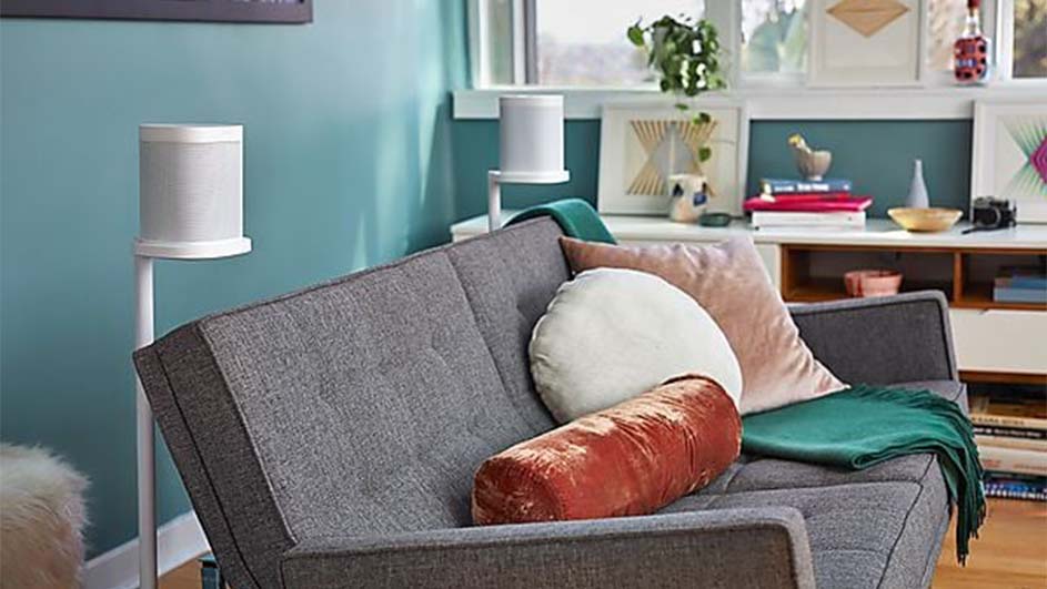 Grijze bank met kussens en groene deken, twee witte speakers op standaards, lichte muren en een kast met decoraties.