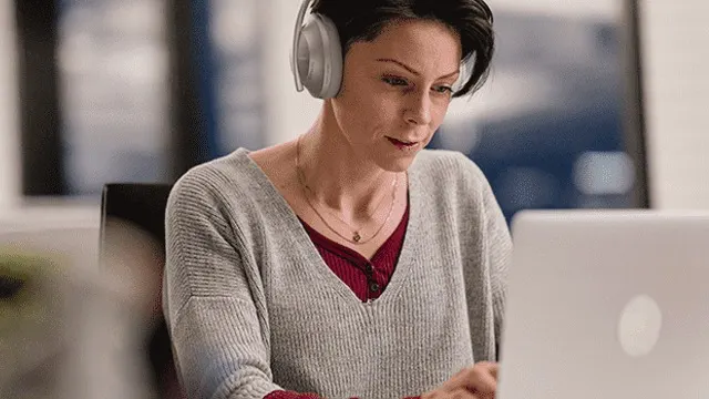 A person wearing gray headphones and a gray sweater works at a laptop. A necklace is visible. The background includes art.