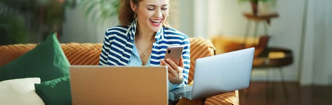 Woman in striped shirt using laptop and smartphone at home, smiling cheerfully, brown box beside her, modern living room.
