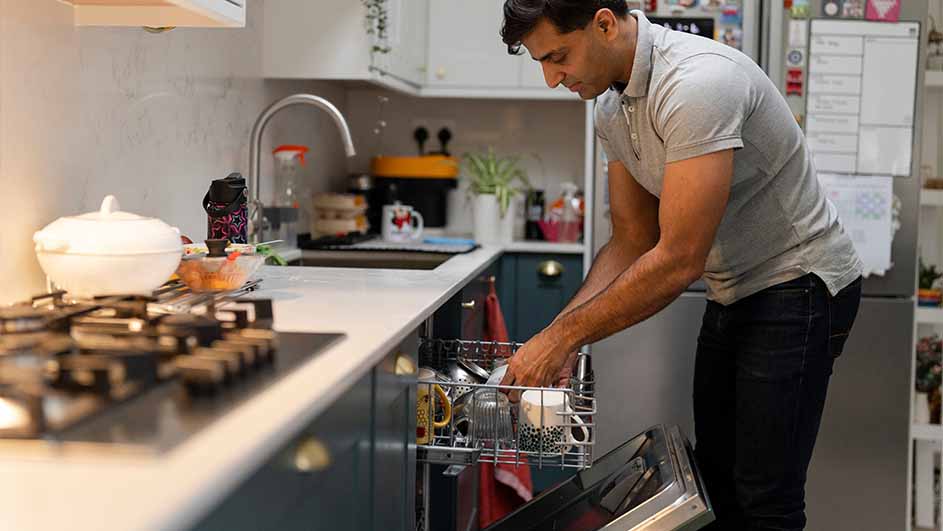 Man laadt vaatwasser in keuken met fornuis, spoelbak en koelkast. Borden en bestek staan in de vaatwasser.