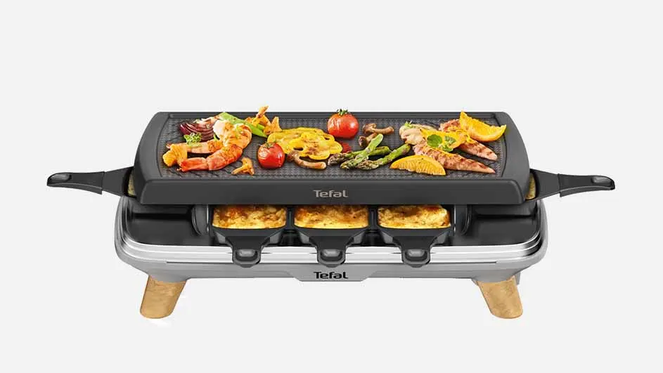 Raclette-grill met garnalen, groenten, kaas in pannetjes. Het apparaat is zwart, grijs en heeft houten poten. Het merk Tefal staat erop.