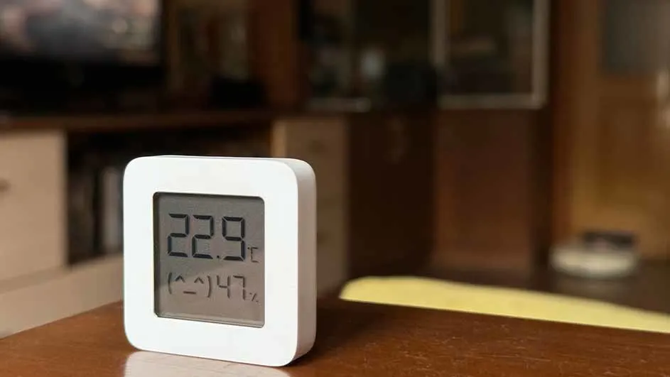 Een witte digitale thermometer toont 22,9 graden Celsius en 47% luchtvochtigheid op een bruine tafel. Achtergrond: meubels.