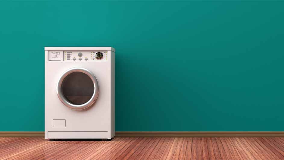 A white washing machine is on a wood floor against a teal wall. The machine has a round door and a control panel on top.