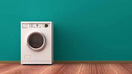A white washing machine is on a wood floor against a teal wall. The machine has a round door and a control panel on top.