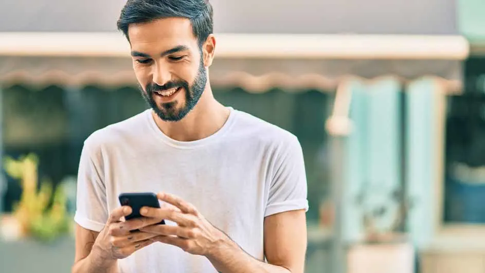 Een glimlachende man met een baard gebruikt een zwarte smartphone buiten, gekleed in een wit T-shirt. Op de achtergrond is een gebouw te zien.