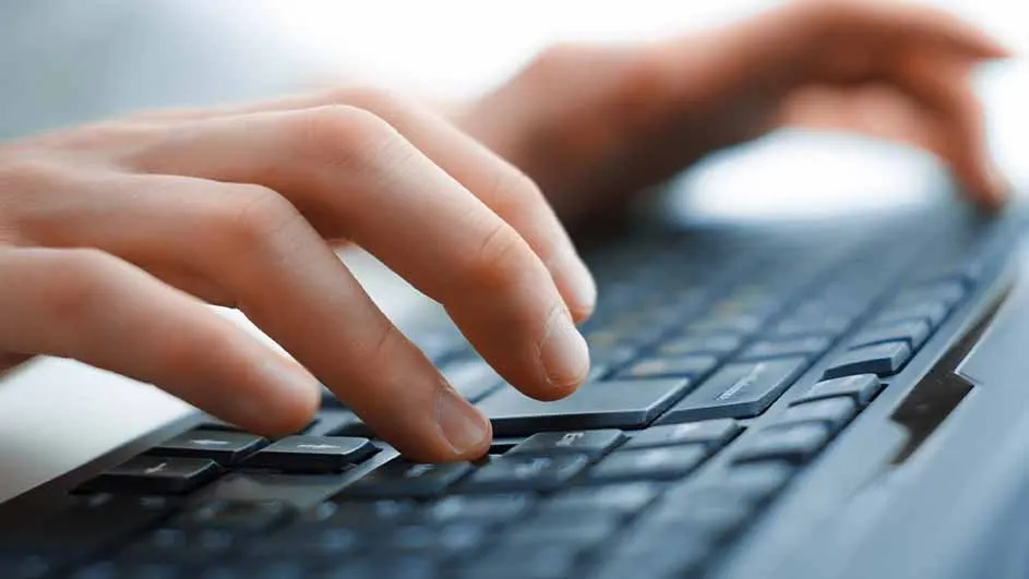Fingers press keys on a dark keyboard. One hand hovers above, ready to type. Background is blurry, light blue.