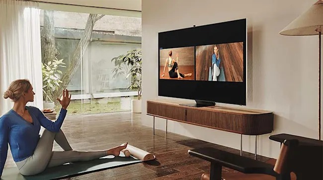 Een vrouw doet yoga-oefeningen op een mat in een lichte woonkamer, terwijl ze een instructievideo volgt op een grote tv aan de muur.