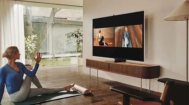 A Samsung TV in a bright living room displays a fitness video; a woman sits on a yoga mat with a roller, surrounded by modern decor and natural light. Brand: Samsung.