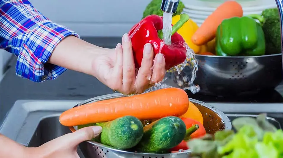 Handen wassen groenten, waaronder rode, gele en groene paprika's, wortelen en komkommers, in een metalen vergiet onder stromend water.