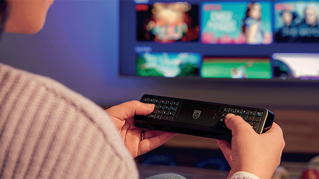 Een persoon gebruikt een zwarte Philips-afstandsbediening voor een tv met diverse beelden. De muur is blauw.