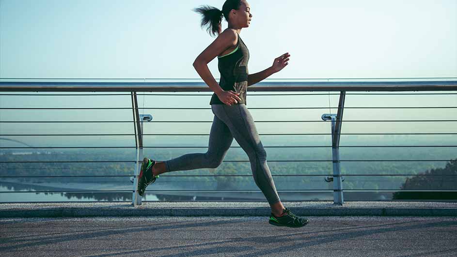 Een vrouw in sportkleding rent op een brug met een metalen reling, met op de achtergrond een rivier en een heldere hemel.