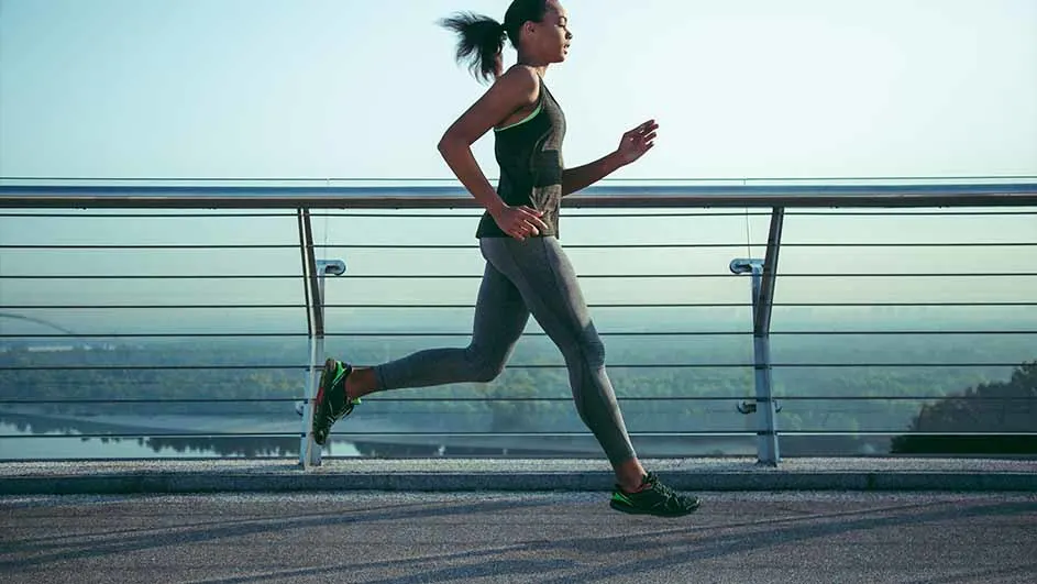 Een vrouw in sportkleding rent op een brug met een metalen reling, met op de achtergrond een rivier en een heldere hemel.