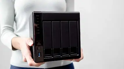 Een persoon houdt een zwarte NAS (Network Attached Storage) apparaat vast tegen een witte achtergrond.