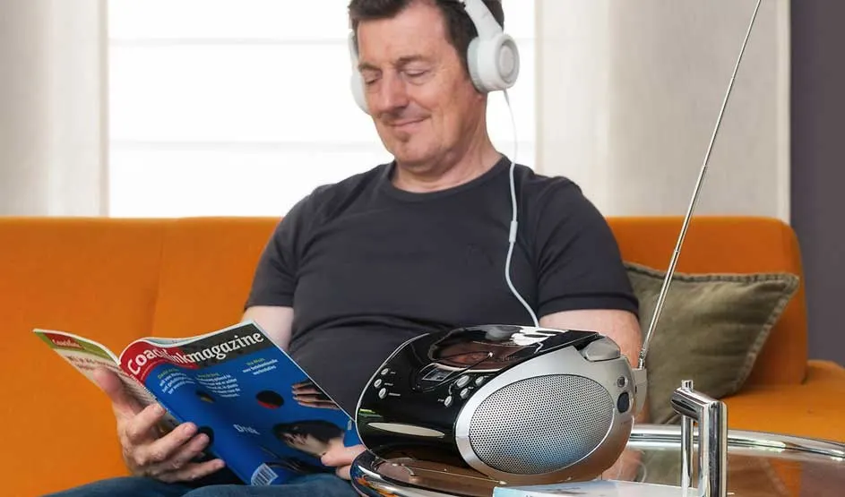 Een persoon zit op een oranje bank, leest een magazine en luistert met witte koptelefoon naar een Philips radio-cd-speler op een glazen tafel.