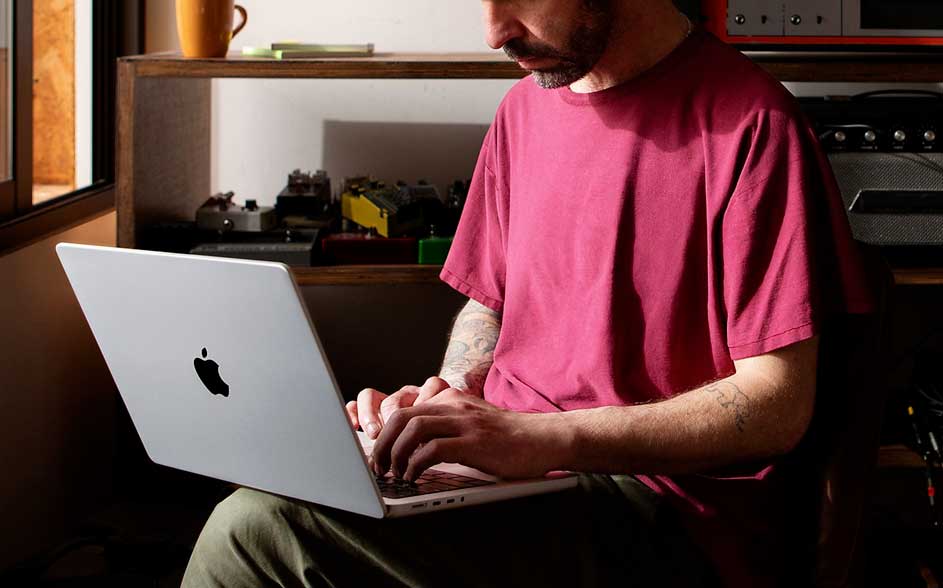 A man in a red T-shirt types on a silver Apple laptop by a window; cups, gadgets and cables sit on the shelf behind.