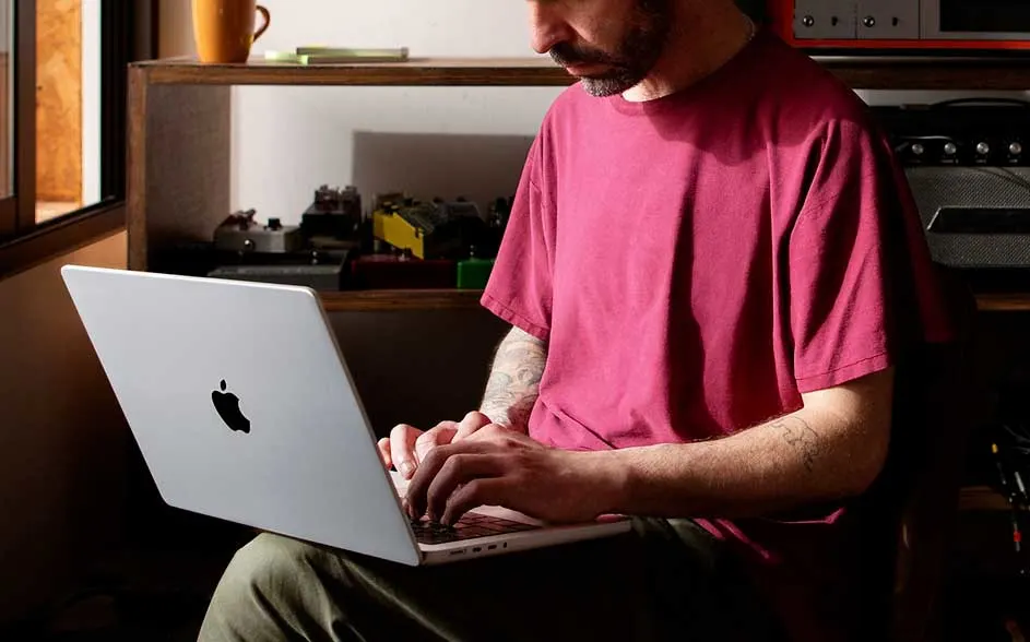A man in a red T-shirt types on a silver Apple laptop by a window; cups, gadgets and cables sit on the shelf behind.