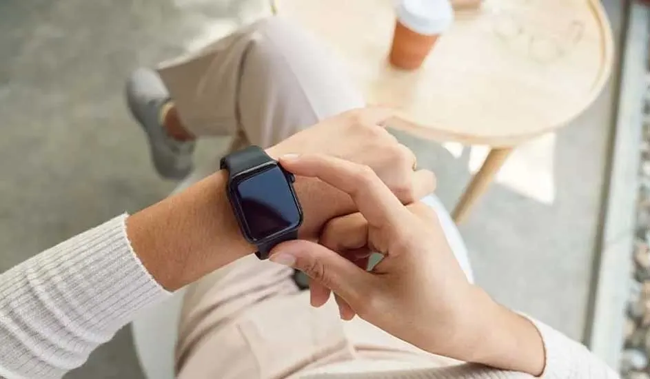 Person in light clothes checking a black smartwatch with blank screen, seated by a small round table with a coffee.