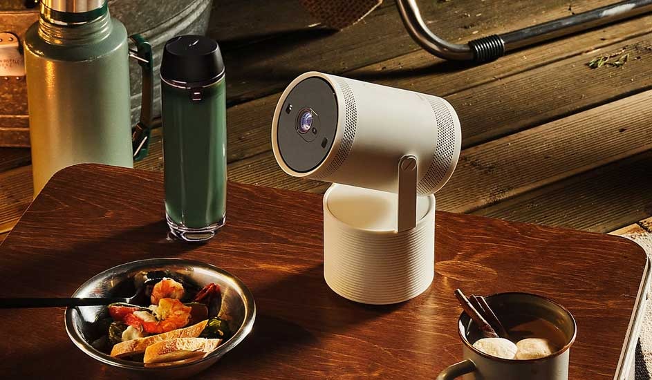 A portable Samsung projector sits on a wooden table outdoors. Nearby are a bowl of food, a mug, a green thermos and camping gear.