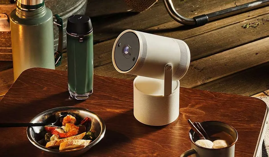 A portable Samsung projector sits on a wooden table outdoors. Nearby are a bowl of food, a mug, a green thermos and camping gear.