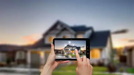 Een tablet met een smart home-interface wordt vastgehouden voor een vervaagd huis bij zonsondergang.