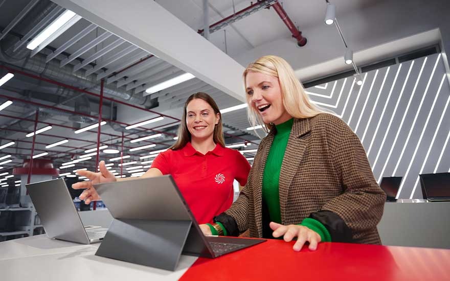 Store employee in red polo helps an excited customer discover a new laptop in a brightly lit, modern electronics shop.
