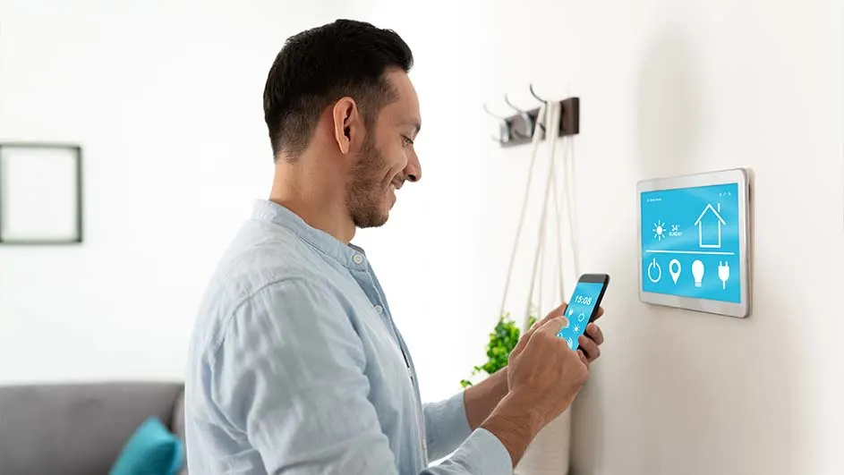 Een man gebruikt een smartphone naast een smart home-bedieningspaneel aan de muur. Haken en een plant zijn zichtbaar.