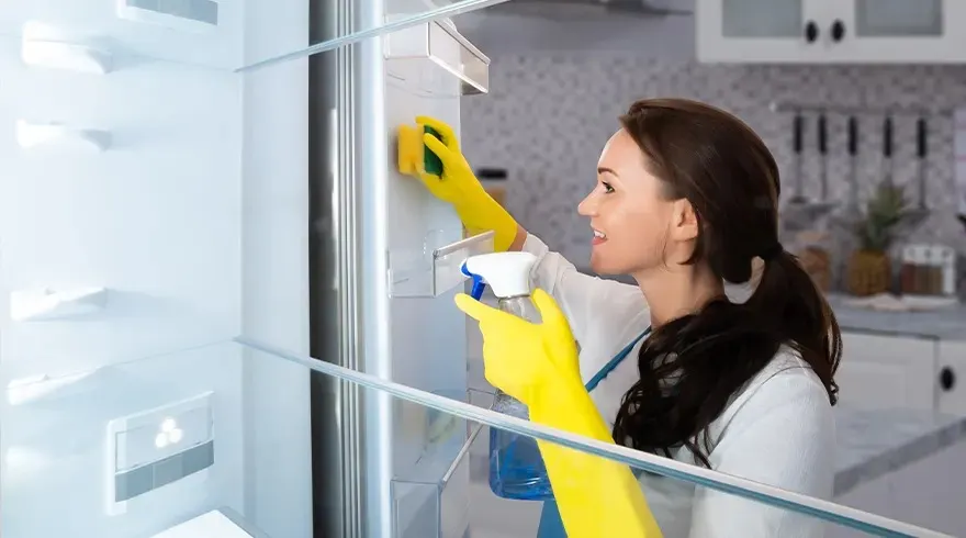 Woman wearing yellow cleaning gloves wipes the inside of a fridge with a sponge and spray bottle in a bright, modern kitchen setting.