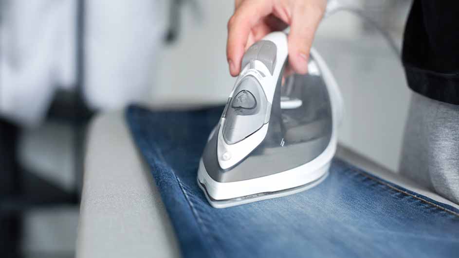 Een hand strijkt een blauwe jeans met een grijs en wit strijkijzer op een wit oppervlak.