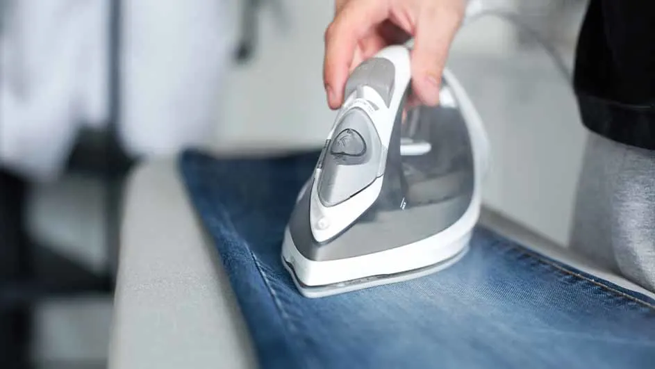 Een hand strijkt een blauwe jeans met een grijs en wit strijkijzer op een wit oppervlak.