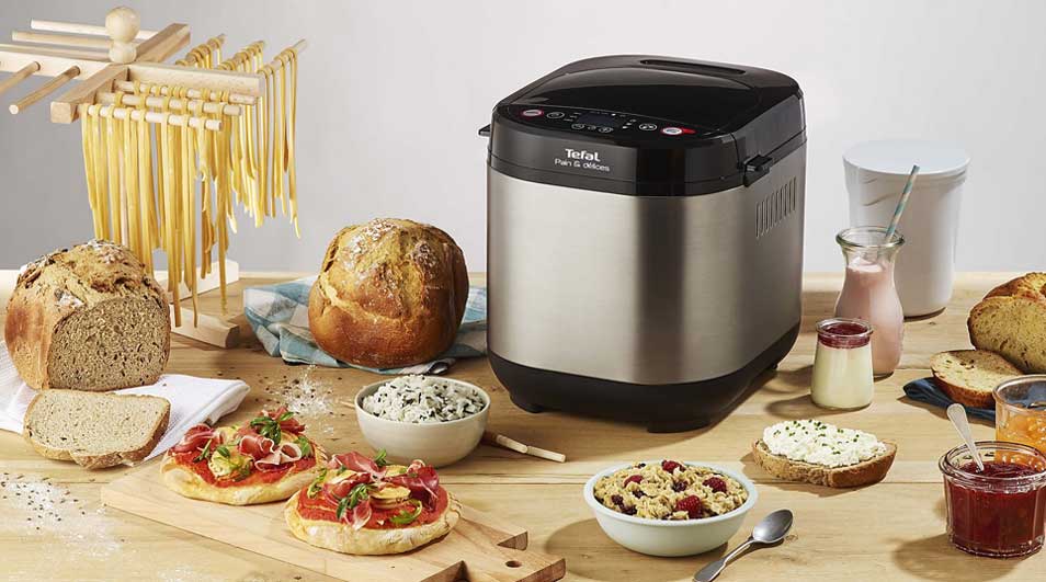 A Tefal bread maker sits on a wooden table, surrounded by fresh loaves, spreads, yoghurt and jars of jam for a generous meal.