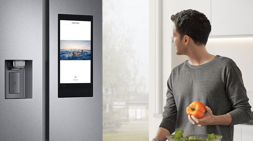 Een man met een paprika naast een grijze koelkast met scherm in een lichte keuken.