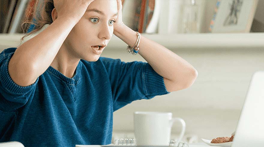 Een vrouw in een blauwe trui kijkt geschrokken naar een laptop. Een witte mok en een koekje staan op de tafel.
