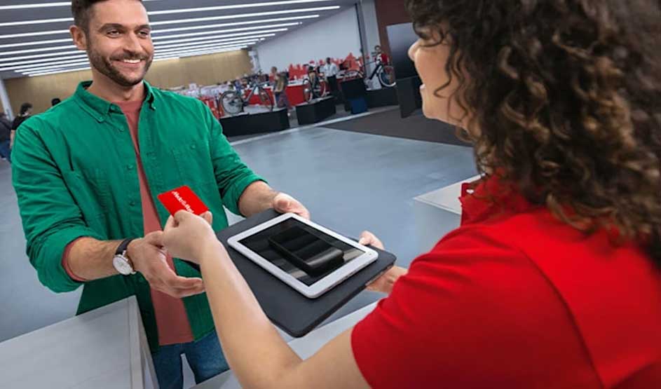 Een klant geeft een rode MediaMarkt-klantenkaart aan een medewerker in rood shirt, die een tablet vasthoudt in een moderne winkel.