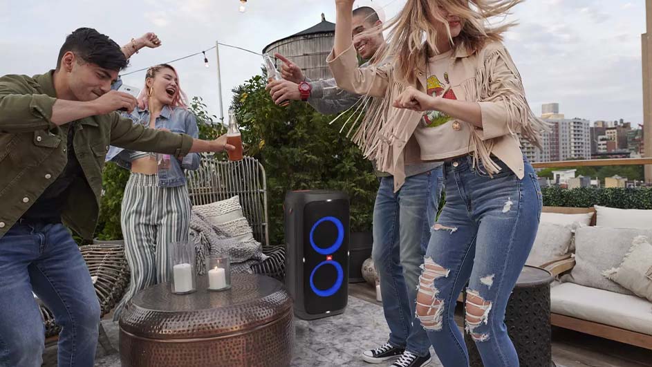 Vier mensen dansen op een dakterras. Een zwarte speaker met blauwe lichten staat centraal, met een tafel en stoelen op de achtergrond.