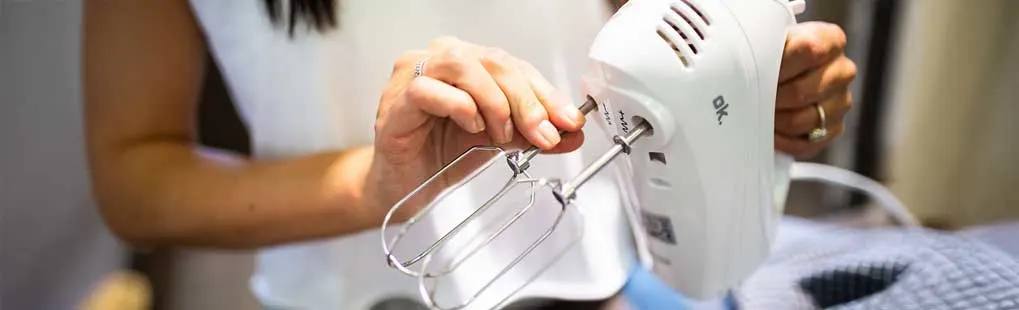 Iemand bevestigt de gardes op een witte handmixer; je ziet handen, metalen kloppers en een aanrecht in zachte, warme keukenverlichting.
